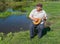 Man having rest on a riverside sitting on a wicker stool, singing and playing mandolin