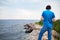 Man with headphones standing with back on rocky grassy seashore in blue t-shirt and jeans