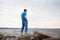 Man with headphones standing in profile on rocky seashore in blue t-shirt and jeans