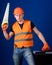 Man in helmet, hard hat carries toolbox and holds handsaw, blue background. Worker, repairer, repairman on serious face