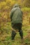 A man in high boots and camouflage stands and urinates in the woods. Rear view. The concept of extreme travel in the taiga