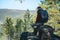 Man hiker with a large backpack sitting on a rock cliff and looking at the sprawling green valley. Freedom in travel