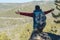 Man hiker with a large backpack stands on a cliff stone in front of a green valley with his arms outstretched