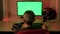 A man in his home office, in front of a green computer screen. Domestic cat walking on the table.