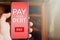 Man holding mobile phone with pay your debt text and pay button on red screen. Online debt payment on smartphone. Financial