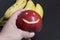 The man is holding a red apple with a carved emoticon in his hand. There is a bananas in the background.