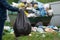 Man holds a full garbage plastic bag in front of overflowing junk container outdoors at countryside