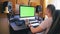 A man in a home music studio sits at a desk with equipment and looks at a green computer screen