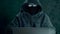 A man in hood sitting at table and coding on laptop. Dark night. Front view close up. Hacker in balaclava mask and