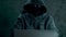 A man in hood sitting at table and coding on laptop. Dark night. Front view close up. Hacker in balaclava mask and