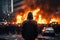 A man in a hooded sweatshirt stands in front of a burning building in Bangkok. Back view Aggressive man without face in hood