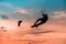 Man kitesurfer athlete jumping at sunset, silhouette at dusk man doing board grab. Kitesurfing sport on orange background