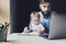 Man and little baby sitting in front of laptop. Serious and busy father with small child on his knees trying to work remotely