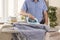 Man in the living room irons the sleeves of a shirt on an ironing stand on an ironing board. In the background, ironed and folde