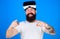 Man with long beard in VR glasses blurred over blue background. Hipster with trendy beard and tattoo pointing at camera