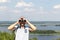 A man looking at camera through binoculars against a scenic view of a wide river on a hill on a sunny day