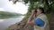 Man looks through binoculars at distance while sitting at scenic river bank on green grass in shadow of tree.