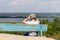 A man looks through binoculars at a scenic view of a wide river while sitting on an old wooden bench on a hill