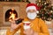 Man in medical mask and santa claus hat, holding cup and bottle with medicines in hands, looks at camera, being ill during New