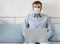 Man in medical mask and shirt works from home in his bed with a laptop