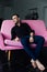 Man model sits on pink sofa in loft interior. Businessman in a black shirt and watch, blue pants.