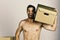 Man with naked torso holding cardboard box on his shoulder. Macho with beard and surprised face carrying box