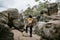 A man in an orange jacket and hat stands on a rock and looks into the distance. Mountain travel concept