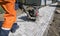 Man in orange uniform using vibrational paving stone machine for finish on a sidewalk road construction site.