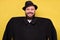 Man in oversize baggy black coat and hat smiling standing on yellow studio