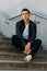 man posing sits on the steps near railing. Handsome young man in stylish black clothes and white shoes posing near a railing