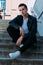 man posing sits on the steps near railing. Handsome young man in stylish black clothes and white shoes posing near a railing