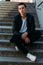 man posing sits on the steps near railing. Handsome young man in stylish black clothes and white shoes posing near a railing