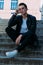 man posing sits on the steps near railing. Handsome young man in stylish black clothes and white shoes posing near a railing