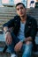 man posing sits on the steps near railing. Handsome young man in stylish black clothes and white shoes watch on hand