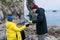 Man pouring hot drink from thermos in a cup for woman. Friends in warm clothes have a rest after walk on rocky seaside