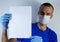Man in a protective mask on a blue background. holds a sheet of white paper vertically. Copyspace for text