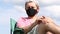 A man in a protective mask and cap sits in a camping chair. Against the background of blue sky. Outdoor recreation in self-