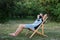 Man in protective medical mask on his face sunbathing outside green lawn in park. Person outdoors relaxing on deck chair in garden