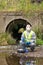 A man in a protective suit and a respirator against bacteria and viruses sits by the river next to a mobile laboratory for