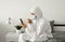 Man in protective white suit and medical mask is using a phone at his home sitting on a bed because of coronavirus