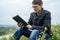 Man reading book in green hilly field. Side view of young handsome man in glasses and warm jacket reading book in open