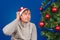 The man in a red cap scratches a nape sitting near a green fir-tree.