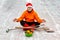 A man in a red Christmas hat and boots, in a orange jacket, sits in a lotus position on the site of his yard with a brush and a