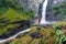 A man with a red hooded jacket stand next to a big waterfall in a national park in Norway