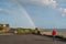 A man in a red jacket walks along a seaside promenade whilst a rainbow reaches down to the sea in front of him