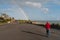 A man in a red jacket walks along a seaside promenade whilst a rainbow reaches down to the sea in front of him