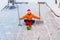 A man in a red New Year hat and orange jacket sits on the site of his yard with a brush and a shovel. He cleaned the snow from the