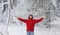 A man in a red sweater stands in a snow-covered forest with his hands in the air. Family winter vacation