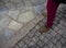 A man in red trousers and brown shoes stands on a tiled and natural stone sidewalk in Greece