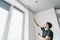 Man with a roller in his hands paints the ceiling in the gray color near the window of his house.